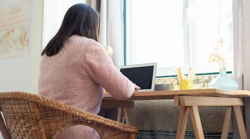 Zdjęcie poglądowe: kobieta widziana od tyłu pracuje przy laptopie przy oknie, siedzi na wiklinowym krześle